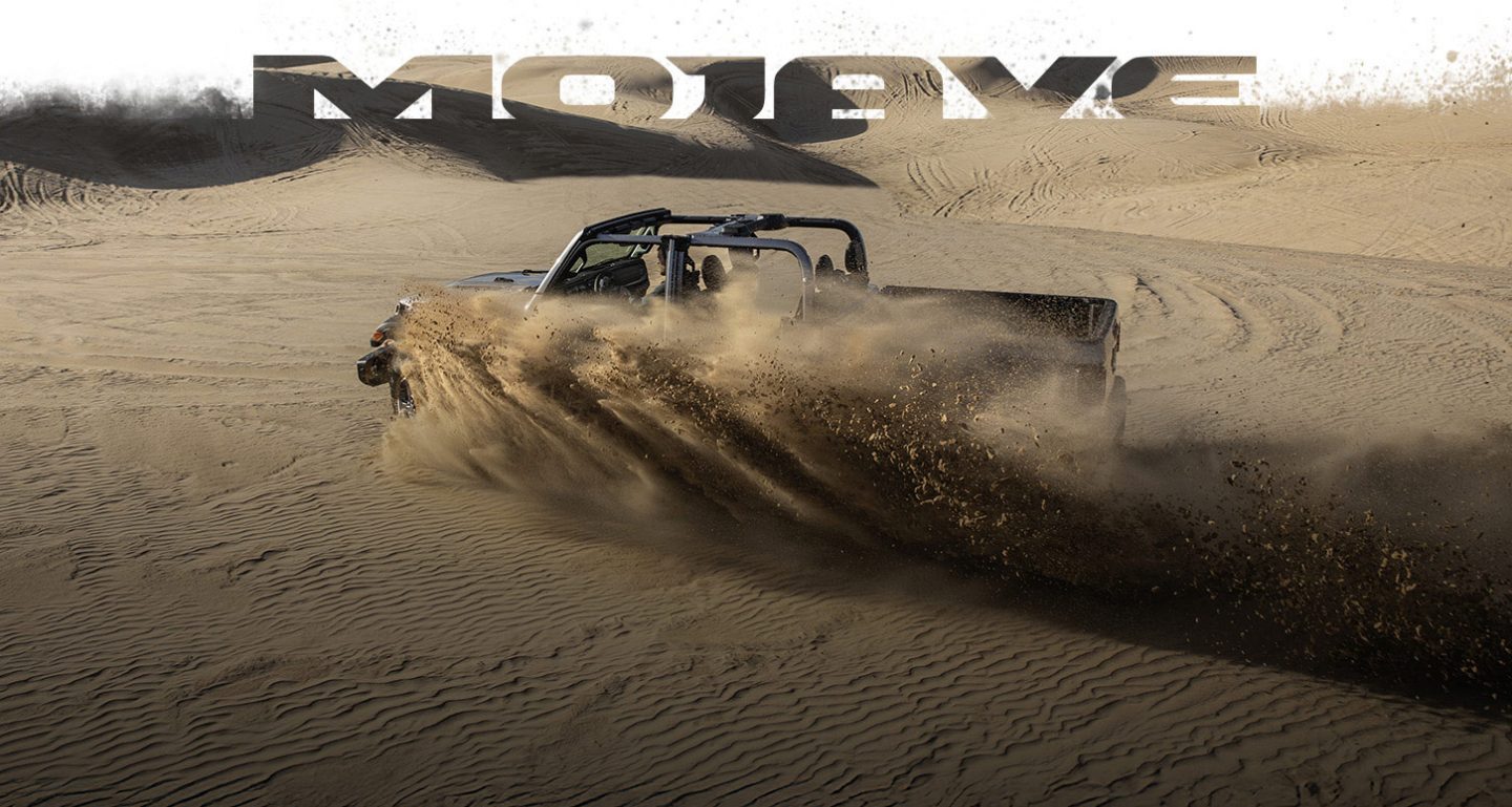 A driver-side profile of a gray 2026 Jeep Gladiator Mojave X with its top removed, traveling through the desert, kicking up sand as it goes. Mojave.