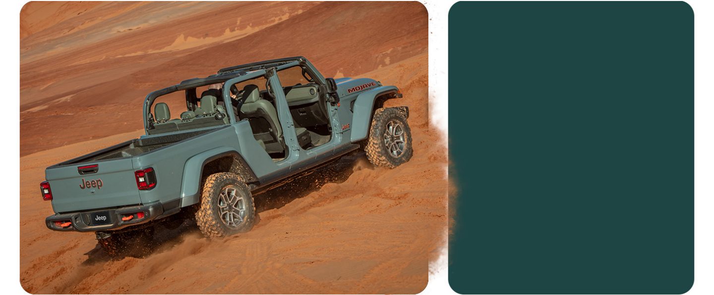 A gray 2026 Jeep Gladiator Mojave X with its doors and top removed, ascending a sand hill in the desert.