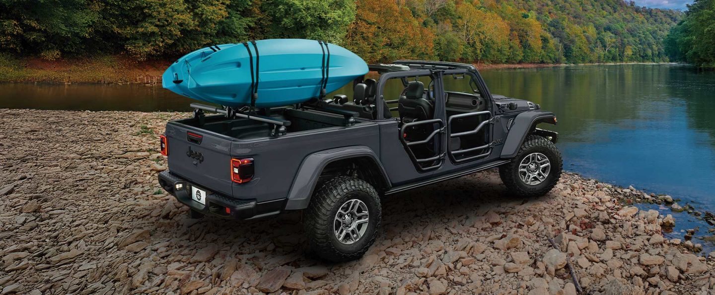 A gray 2026 Jeep Gladiator with tube doors, its top removed and two kayaks strapped to its pickup box mounting system, parked on a rocky beach beside a lake.